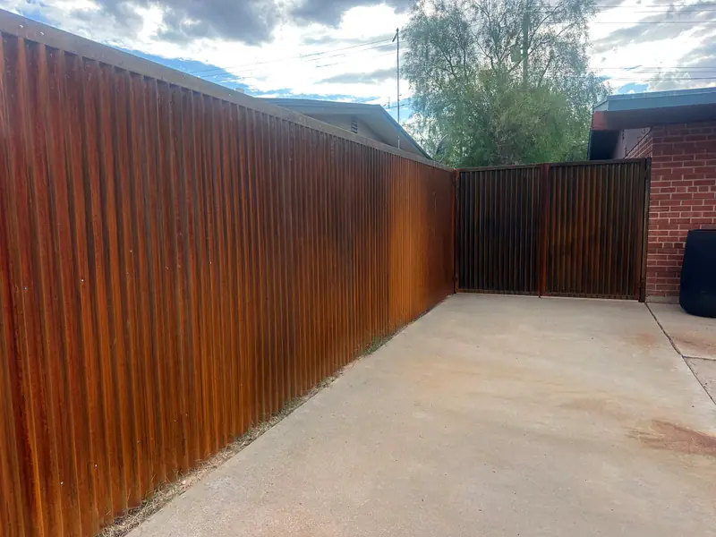 Long run of raw corrugated steel fence with a capped top along a concrete driveway